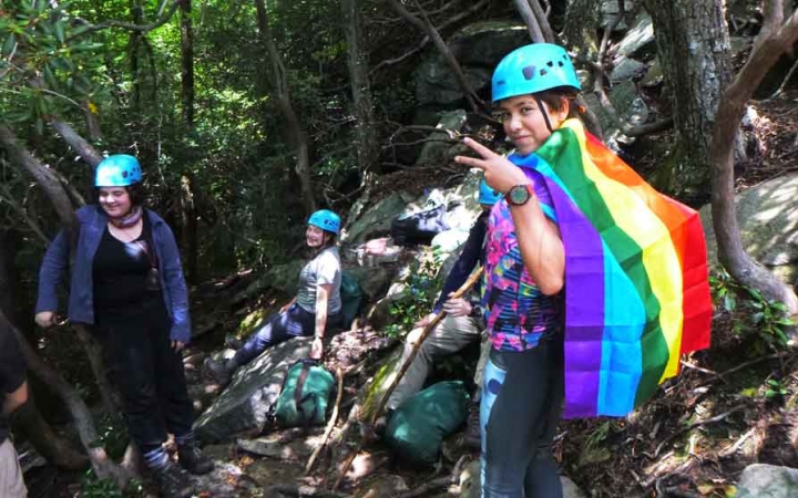 a student wearing a rainbow flag as a cape gives a peace sign while on a rock climbing trip in north carolina